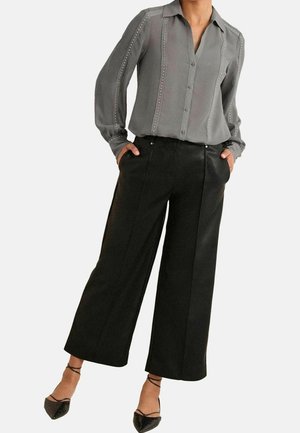 Kvinde iført en grå skjorte med knapper med nittede lodrette striber og sorte vidde bukser med korte ben, hænderne i lommerne, sorte spidse flade sko.