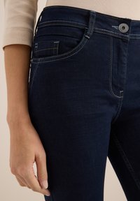 Donkerblauwe denim jeans met een slim fit, vijf-pocket ontwerp, lichte stiksels en een knoopsluiting bij de taille. Zachte textuur is zichtbaar.