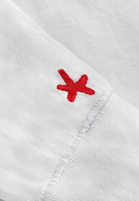 Biała tkanina z drobnym prążkowanym wzorem, z małym czerwonym haftowanym akcentem w kształcie gwiazdki przy krawędzi mankietu.