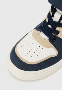 Laivastonsininen ja beige mokkakengät, joissa on rei'itetty valkoinen varvastoppa, litteät laivastonsiniset nauhat ja kumipohja. Varustettu pehmustetulla nilkkakauluksella.