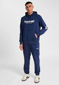 Marinblå hoodie och sweatpants i mjukt material med framficka och logotryck. Detaljer med vita ränder på ärmarna.