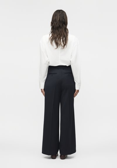 Γυναίκα με μακριά κυματιστά μαλλιά, που φοράει λευκό πουκάμισο με μακριά μανίκια και μαύρο παντελόνι με ψηλή μέση και φαρδύ πόδι, στέκεται με την πλάτη γυρισμένη.