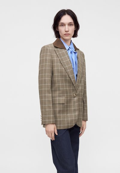 Personne portant un blazer à carreaux marron avec un col foncé sur un chemisier bleu avec un nœud noué, associé à un pantalon foncé, debout devant un fond blanc.