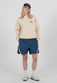 Person steht mit den Händen in den Taschen, trägt einen beigen Pullover, blaue Shorts, weiße Socken, Loafers und eine blau-schwarze Schirmmütze.
