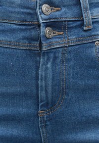 Denim jeans met een donkerblauwe was, voorzien van twee metalen knopen in de taille, contrasterende gouden stiksels en een voorzak.