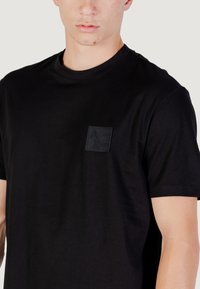Camiseta de algodón negra con cuello redondo, que presenta un parche de logo texturizado negro en la parte izquierda del pecho. Mangas cortas.