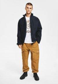 Granatowa kurtka zapinana na zamek błyskawiczny z kieszeniami, biała koszulka z pomarańczową grafiką oraz brązowe spodnie cargo z zamkami błyskawicznymi. Czarnie buty trekkingowe dopełniają strój.