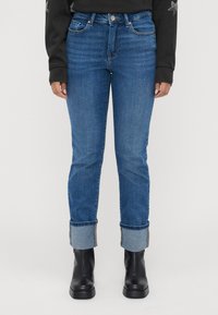 Person med blå oppbrettede jeans, svarte lær ankelstøvler og en svart langermet topp med stjernedesign på ermene.