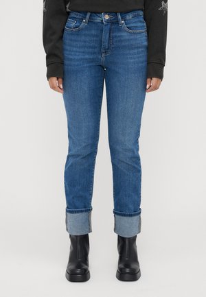 Person med blå oppbrettede jeans, svarte lær ankelstøvler og en svart langermet topp med stjernedesign på ermene.