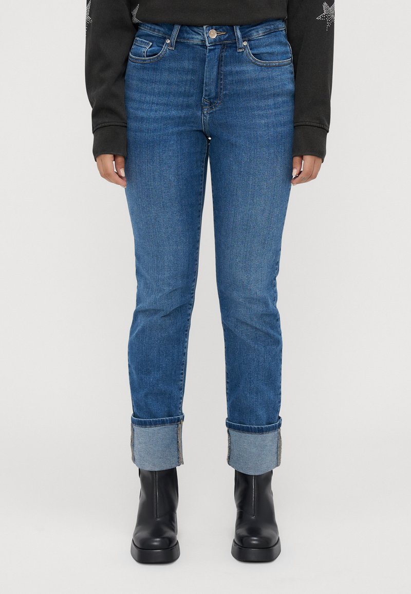 Person med blå oppbrettede jeans, svarte lær ankelstøvler og en svart langermet topp med stjernedesign på ermene.