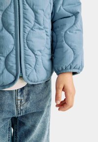 Ljusblå quiltad jacka med dragkedja, prydd med vågiga sömmar och elastiska ärmslut, i kombination med blå denimjeans.