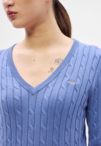 Gaiši zils adīts džemperis ar V veida izgriezumu, ar teksturētām rakstu niansēm un mazu logotipa akcentu uz krūtīm. Mīksts, adīts audums.