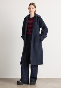 Cappotto lungo in lana di colore blu scuro con un ampio colletto, chiusura con bottoni e tasche frontali. Abbinato a un maglione a righe rosso e blu navy e pantaloni scuri.