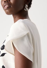 Top bianco con texture e maniche corte a campana, decorato con un motivo di foglie nere. Orecchini circolari lucidi completano l'outfit.