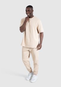 Beige katoenen t-shirt en bijpassende joggers, ruime pasvorm, korte mouwen, met logo op de borst, gecombineerd met witte sport schoenen.