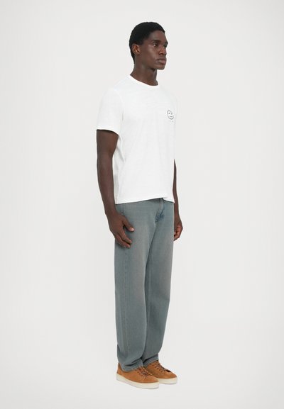 White short-sleeve t-shirt with a round neckline and a printed smiley face on the chest, paired with light grey loose-fit denim jeans and brown sneakers.