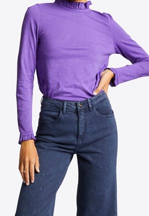 Mujer con una blusa de manga larga morada con cuello y puños con volantes, combinada con vaqueros de pierna ancha de talle alto en azul oscuro.