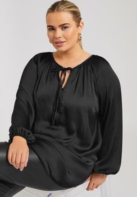 Svart satinblus med långa ärmar, rynkad halsringning, knytdetalj och knapplist. Texturen ser slät ut med en subtil lyster.