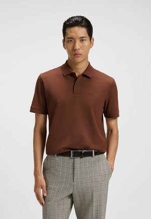 Polo marrone con colletto classico, maniche corte e un logo discreto sul petto, abbinato a pantaloni a quadri e una cintura nera.