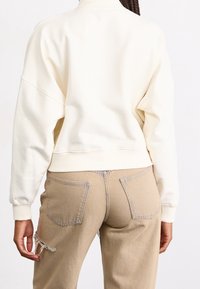 Felpa crema con spalle cadute e polsini a costine, abbinata a pantaloni beige con orlo sfilacciato e tasche posteriori.