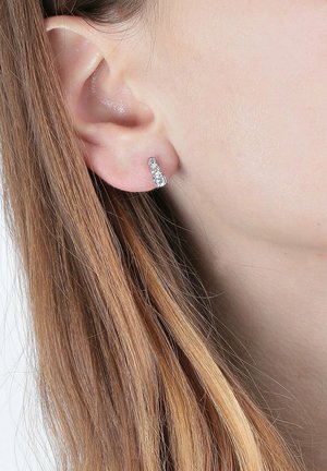 Sølv øreringe har et geometrisk design med en struktureret overflade, placeret tæt på øret. Øreflippen er synlig og viser hår i nærheden.