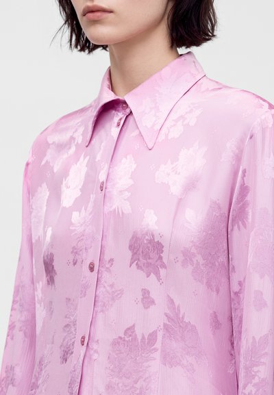 Femme portant un chemisier rose transparent boutonné avec un motif floral et un col pointu, visible du cou jusqu'au milieu du torse.