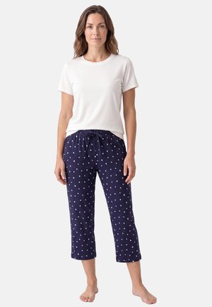 Femme debout pieds nus portant une chemise blanche à manches courtes et un pantalon court bleu marine avec de petits motifs en forme de cœur blancs.