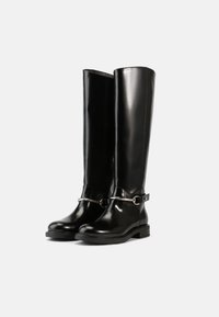 Botas pretas até ao joelho, feitas de material brilhante, com um detalhe de fita e anel prateados. A sola grossa preta oferece estabilidade e durabilidade.