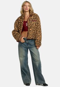 Korte, tan en zwart pattered fleecejack met hoge kraag en zak met rits, gecombineerd met losse blauwe denim jeans en zwarte schoenen.