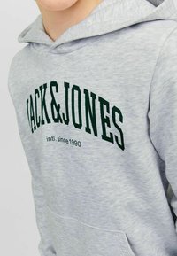 Sudadera gris con bolsillo frontal, con texto verde oscuro "JACK&JONES" y "intl. desde 1990" en el pecho. Material suave de mezcla de algodón.