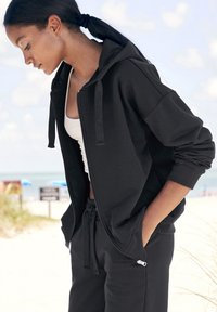 Sudadera negra con cremallera y capucha ajustable, de corte relajado, con puños acanalados y bolsillos, combinada con pantalones de chándal negros a juego, en un entorno al aire libre.