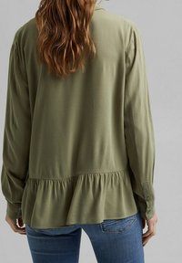 Blusa lunga a maniche con verde oliva, con orlo arricciato; realizzata in tessuto morbido. Presenta polsini con bottoni e una vestibilità comoda, con una texture liscia.