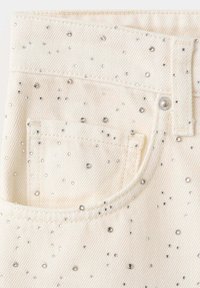 Gros plan sur un pantalon en tissu crème avec des clous argentés dispersés, montrant la taille, la boucle de ceinture, le bouton et les détails de la poche avant.