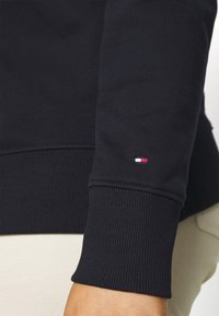 Felpa navy con polsini a costine, caratterizzata da un piccolo logo rosso, bianco e blu sulla manica. Texture liscia. Primo piano sul braccio.
