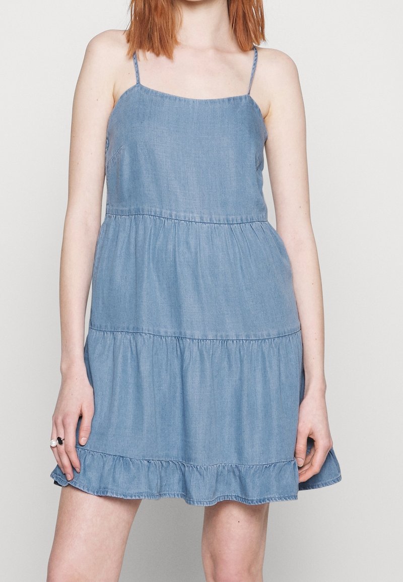 Robe en jean à fines bretelles, de couleur bleu clair, ourlet volanté à volants en étages, et coupe décontractée. Fabriquée en matériau en jean doux avec une texture lisse.