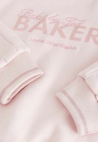 Gaiši rozā džemperis ar ribotām aprocēm. Uzraksts "Baker by Ted BAKER" tumšākā rozā krāsā, blakus "Londona, Apvienotā Karaliste". Mīksta tekstūra.