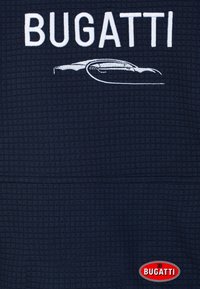 Logo Bugatti e sagoma dell'auto ricamati in bianco su un tessuto scuro e strutturato, con una toppa ovale rossa Bugatti sottostante.