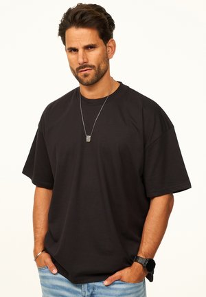 Mann mit dunklem Haar und Bart, der ein schwarzes, übergroßes T-Shirt, hellblaue Jeans, eine silberne Halskette, eine Uhr und ein Armband trägt und mit den Händen in den Taschen steht.