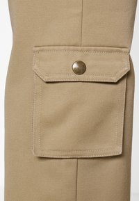 Beige cargo-housut, joissa on teksturoitu pinta. Varustettu sivutaskulla, jossa on läppä ja metallinen nepparikiinnitys. Selkeä ompelusauma on näkyvissä.