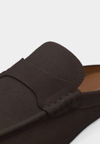 Κοντινό πλάνο ενός μοκασίνι από σκούρο καφέ δέρμα σουέτ με ραμμένο λουράκι και πλεκτή λεπτομέρεια στο πάνω μέρος.