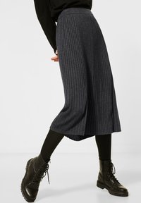 Jupe plissée gris foncé à taille haute, en tissu texturé. Associée à des bottes de combat noires à lacets et à des leggings noirs.