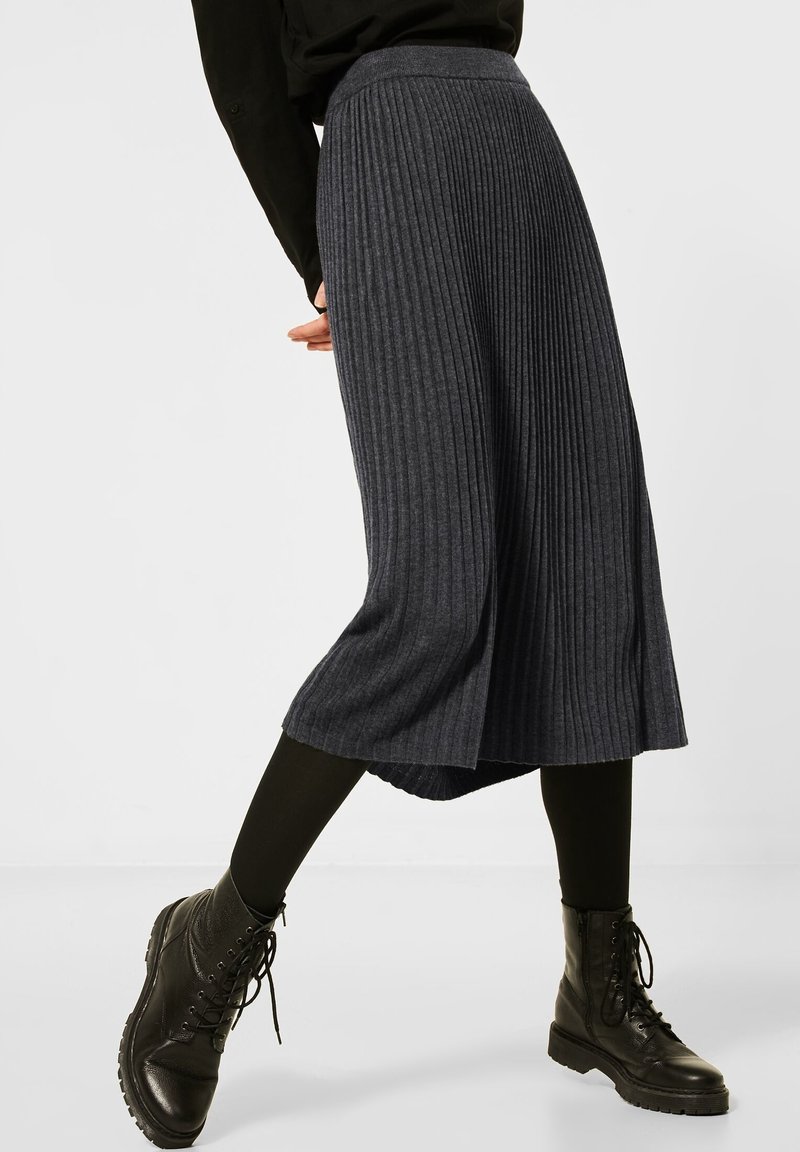 Jupe plissée gris foncé à taille haute, en tissu texturé. Associée à des bottes de combat noires à lacets et à des leggings noirs.