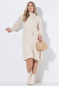 Abito lavorato a maglia crema con collo alto e dettaglio in vita legato, abbinato a una borsa a tracolla beige intrecciata e stivaletti crema, con una texture complessivamente liscia.