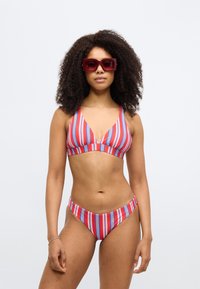 Piros és kék csíkos bikini szett V-nyakú felsővel és alacsony derekú alsóval. Fényes piros, téglalap alakú napszemüveg emeli ki a megjelenést. Sima anyagtextúra.
