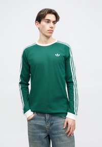 Zelená košeľa s dlhým rukávom a bielym golierom a manžetami, s bielymi trojpruhovými akcentmi na rukávoch a malým potlačeným logom na hrudi.