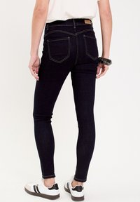 Jean slim en denim bleu foncé avec des surpiqûres jaunes, deux poches arrière et une étiquette en cuir à la taille. Porté avec des baskets blanches.
