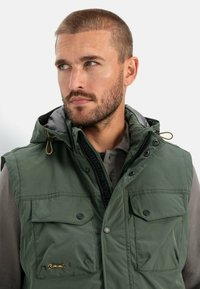 Grüne gepolsterte Weste mit Kapuze, ausgestattet mit zwei Brusttaschen, Druckknopfverschlüssen und einem kontrastierenden Innenfutter. Strukturiert Stoff mit sichtbaren Nähten.