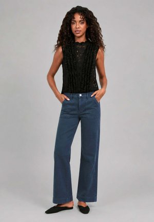 Femme aux cheveux bouclés portant un top sans manches en dentelle noire, un pantalon bleu taille haute à jambes larges, et des chaussures plates noires, debout les mains dans les poches.