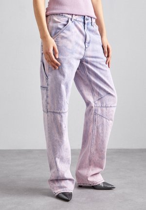 Osoba nosząca luźne, kwasowo-myty różowo-fioletowe dżinsy ze szwami oraz czarne buty z szpiczastym noskiem, stojąca na szarej podłodze.