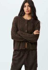 Brauner gestrickter Cardigan mit Kabelmustern, gerippten Bündchen und einer knopfgeschlossenen Front. Kombiniert mit schwarzen Jogginghosen mit Kordelzug.